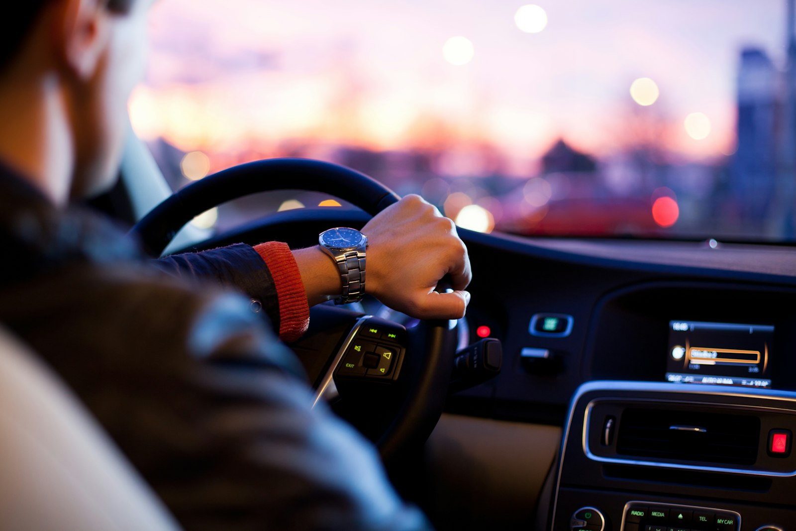 Driver's hand on the steering wheel of a clean car interior at dusk, city lights beyond the windscreen