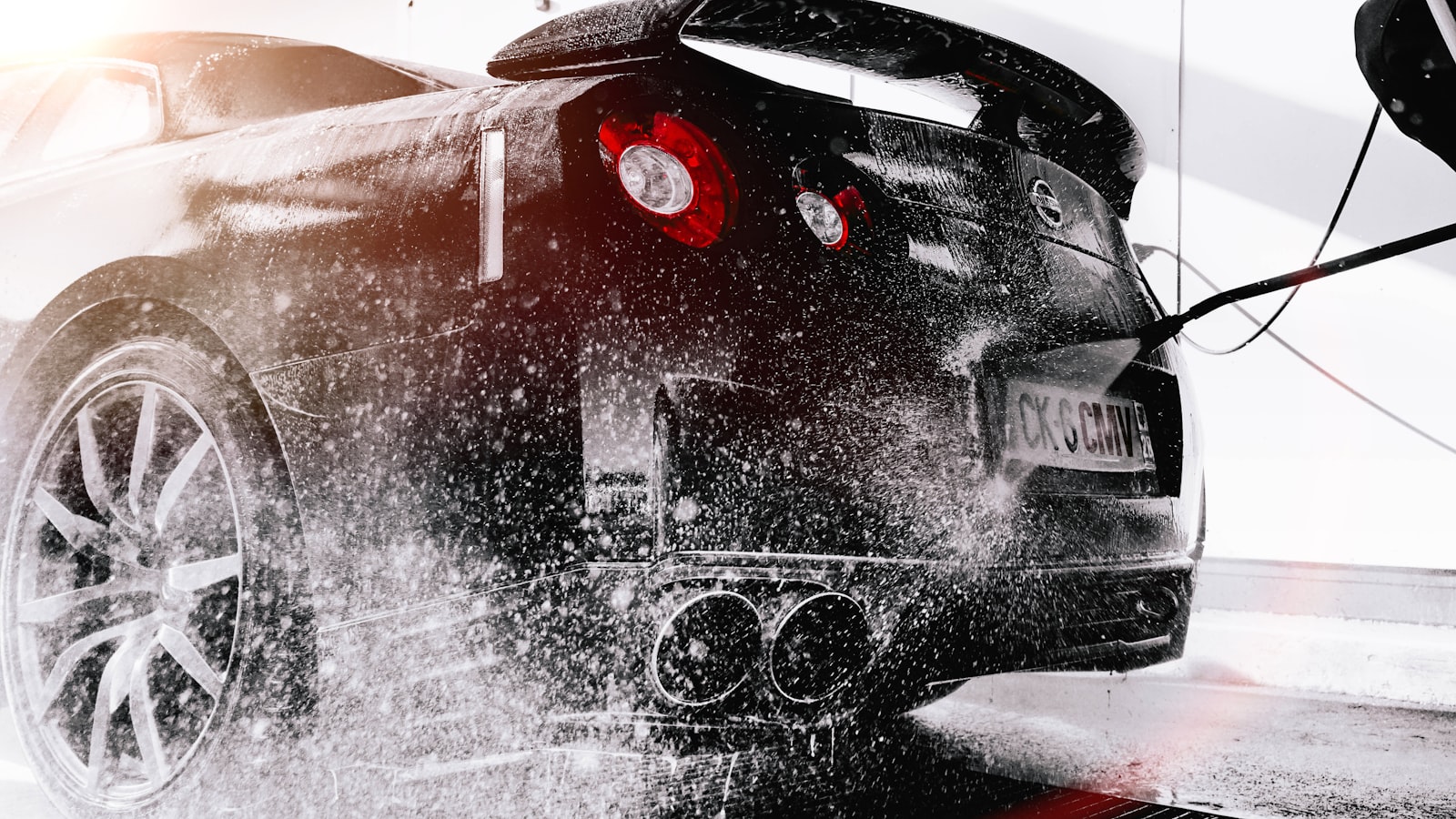 A dark sports car being rinsed with a pressure washer, spray catching the light