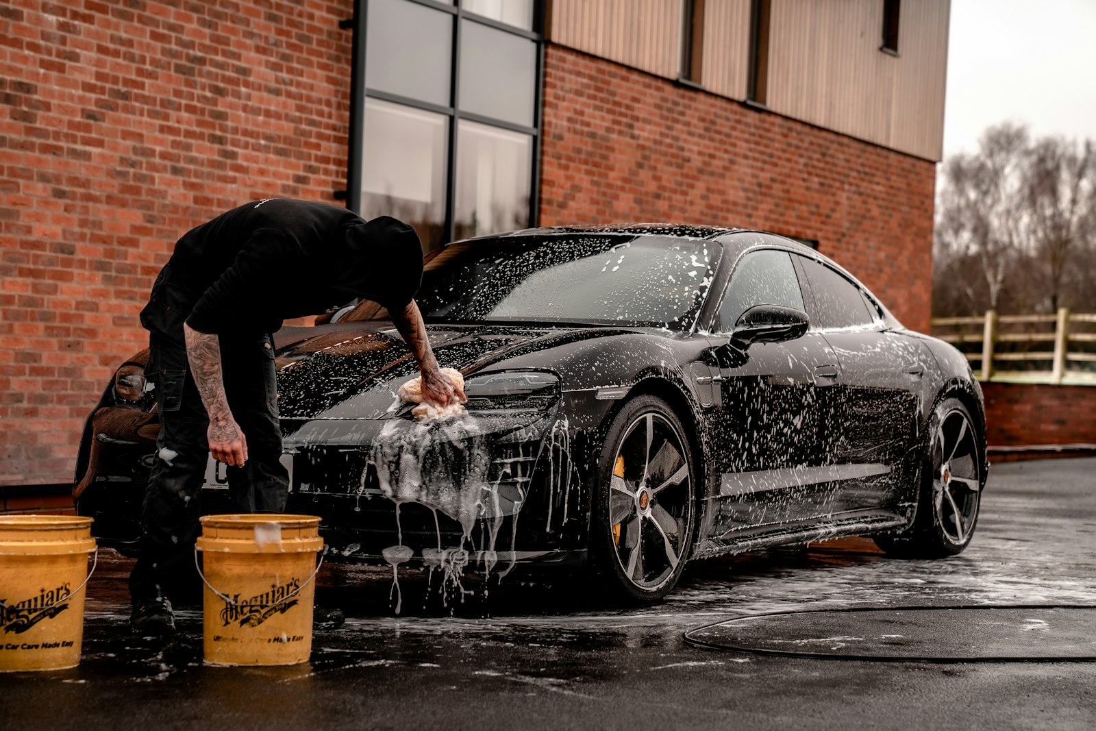 A detailer washing a black sports car with a foam-soaked mitt and two wash buckets